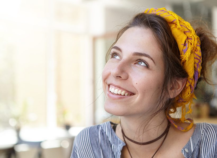 Casual cheerful woman with yellow headband, dark hair and brown eyes sitting at home looking up having shining smile having thoughts admiring summer and her vacations. Human facial expressions concept